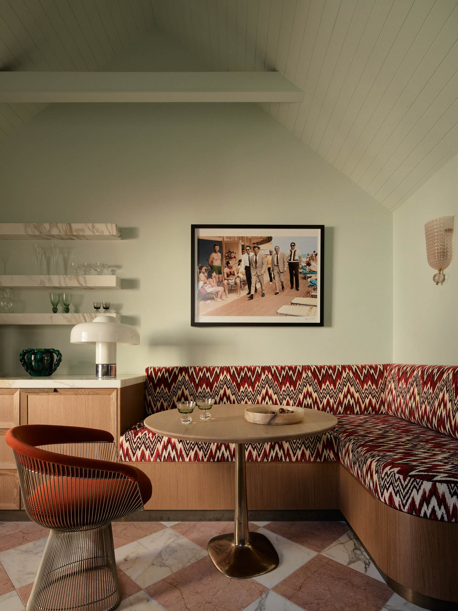 Small dining nook with a round table, patterned bench seating, an orange chair, wall shelves with glasses, and a framed photo on a pale green wall under a slanted ceiling.