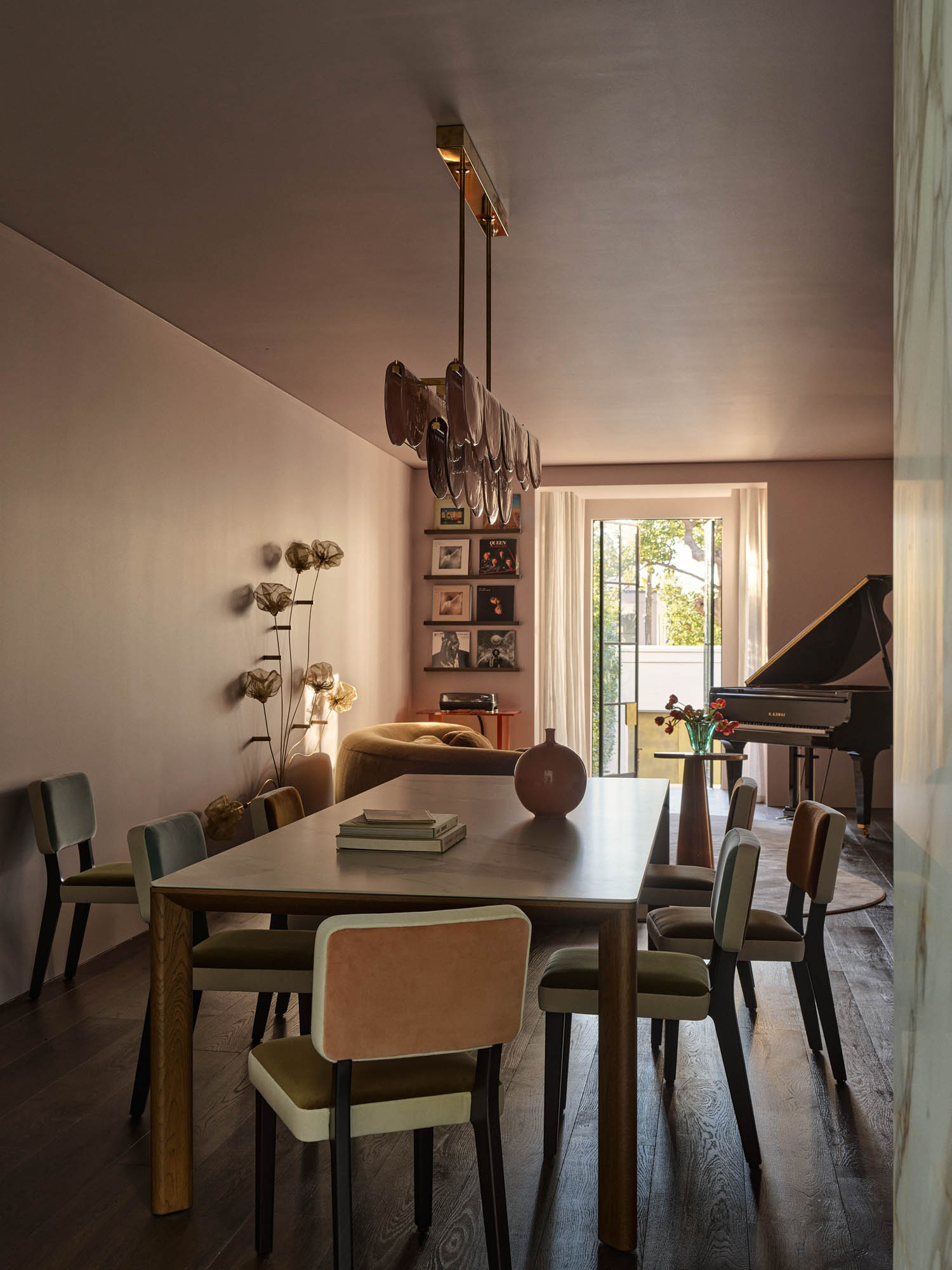 A modern dining room with a wooden table, colorful chairs, a decorative light fixture, wall shelves, floor lamp, and a grand piano near a window with sheer curtains.