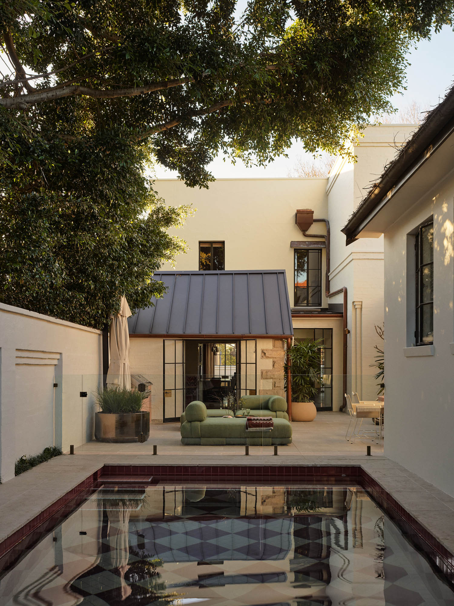 A modern patio area with a green sofa, plants, and a geometric-tiled reflecting pool in front of a two-story cream-colored house.