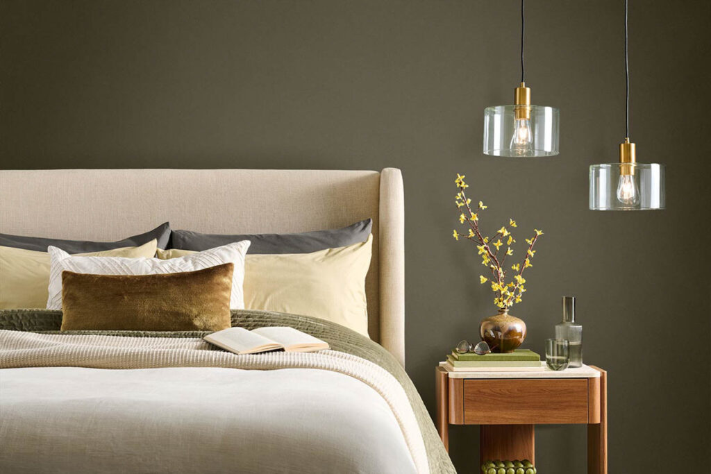 A neatly made bed with neutral bedding sits beside a wooden nightstand with a vase of yellow flowers, a book, and two hanging pendant lights against an olive green wall.