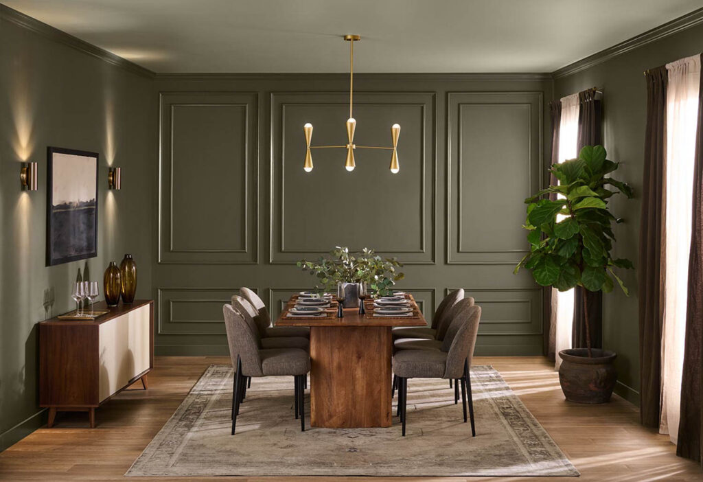 Modern dining room with a wooden table, eight chairs, sideboard, pendant light, large plant, and neutral decor in olive green tones. Natural light enters through window with sheer curtains.