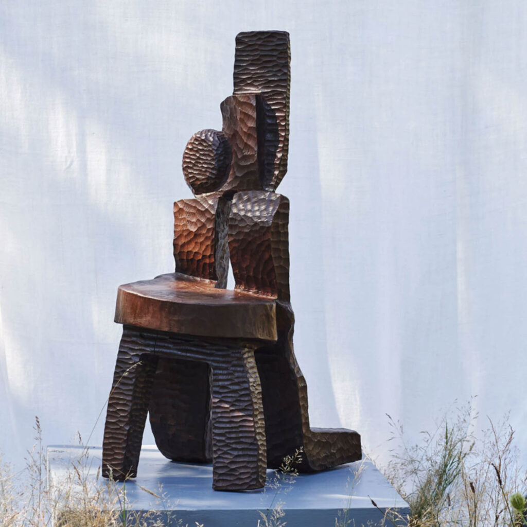 Abstract wooden sculpture with textured, carved surfaces stands on a platform outdoors, set against a white fabric backdrop with grass in the foreground.