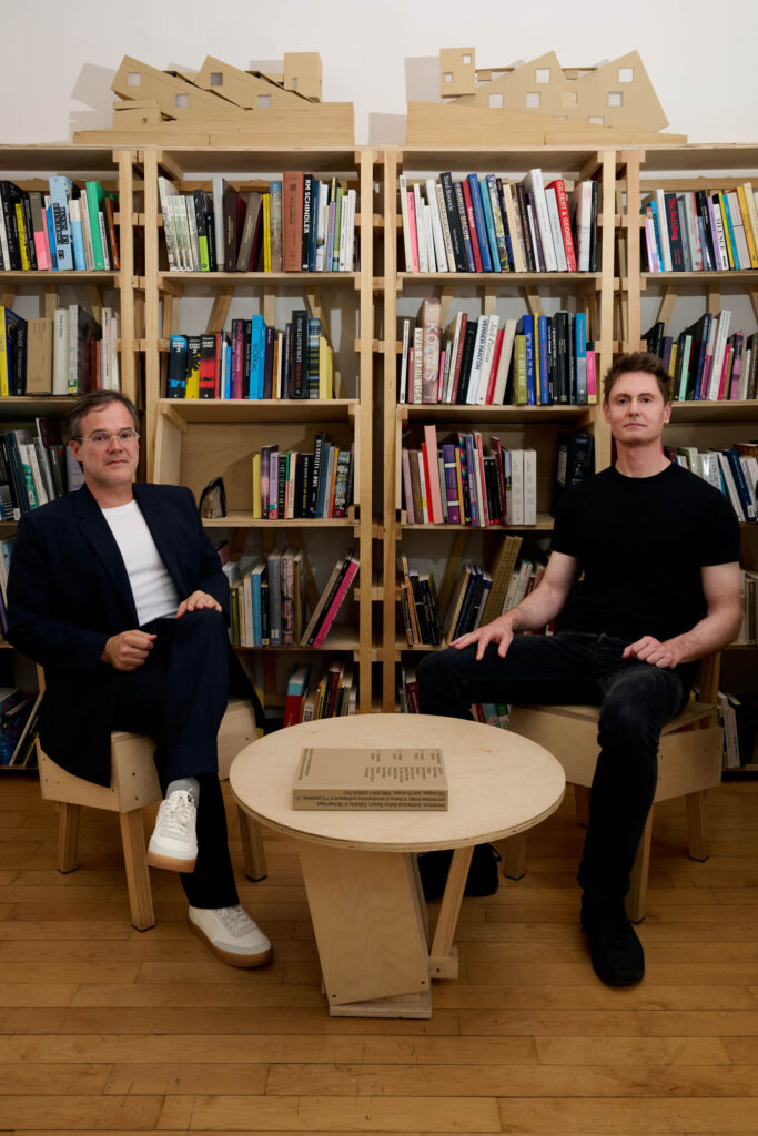 Two men sit on wooden chairs in front of bookshelves filled with books, with a round wooden table and a closed book between them. Model buildings are displayed on top of the shelves.