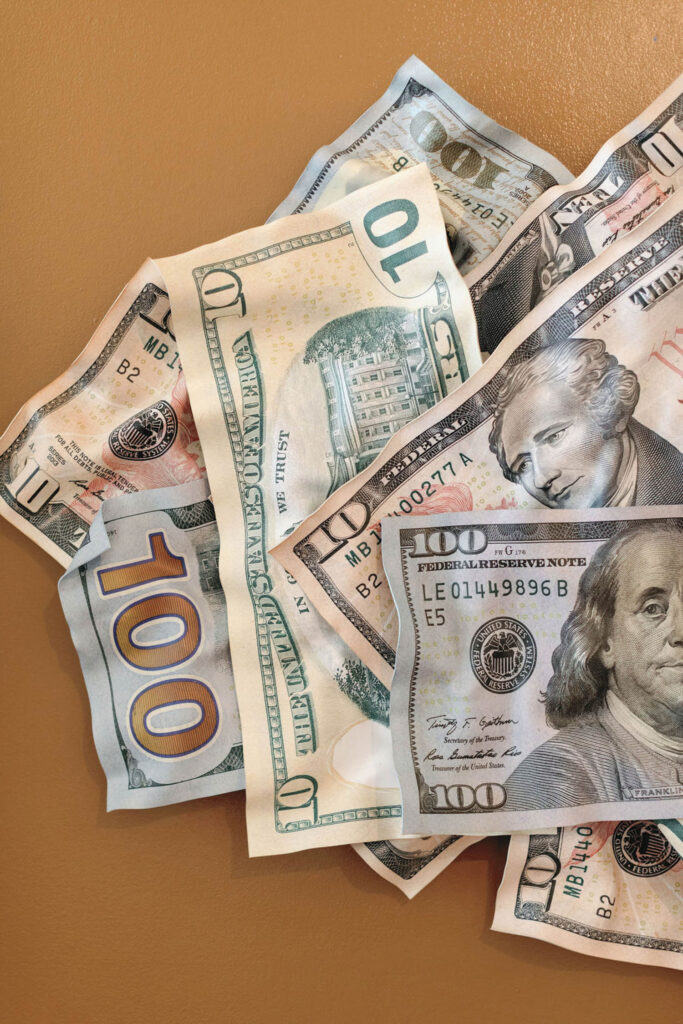 A pile of U.S. dollar bills, including $10 and $100 notes, spread out on a brown surface.