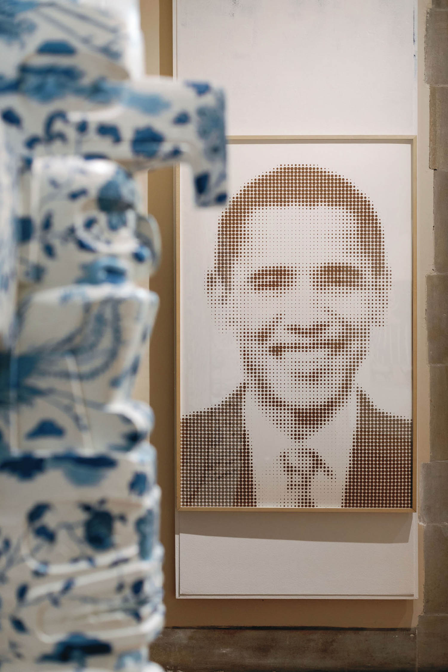 A dot-pattern portrait of a smiling man in a suit and tie is displayed on a wall, partially obscured by a blue and white patterned object in the foreground.