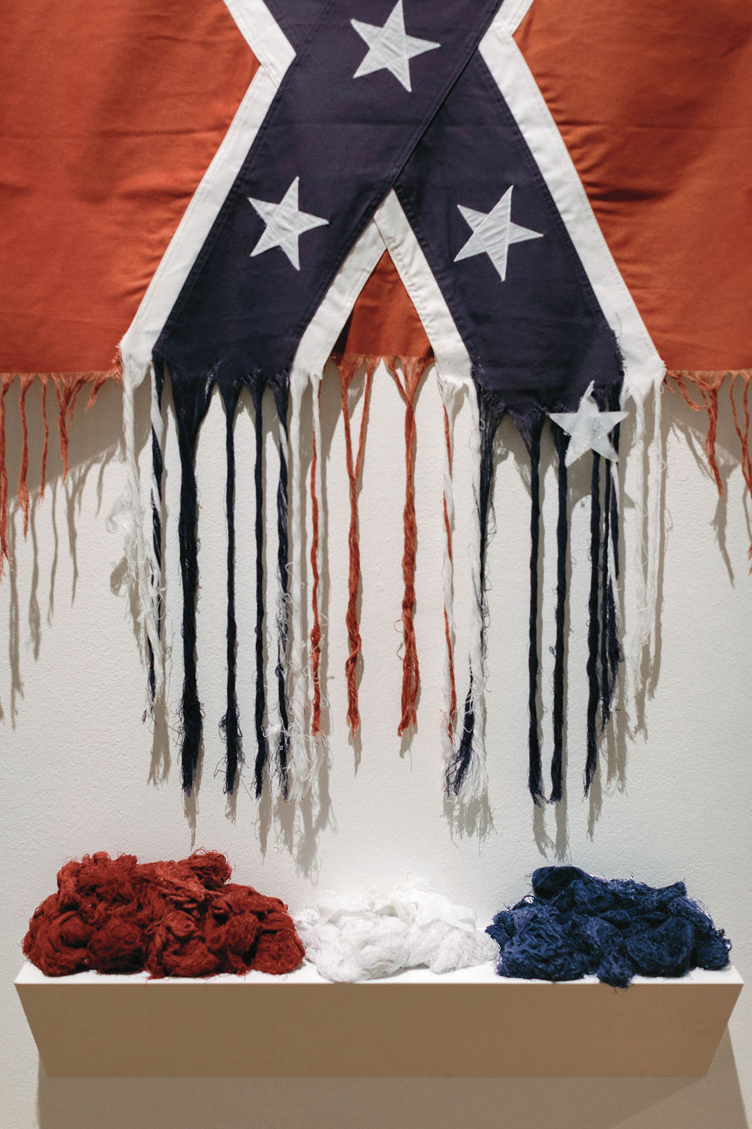 A tattered Confederate flag hangs above a shelf with three piles of torn fabric in red, white, and blue.