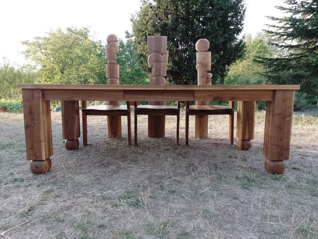 A large wooden table with four chairs is placed outdoors on dry grass, featuring abstract wooden sculptures on top. Tall trees and greenery are visible in the background.