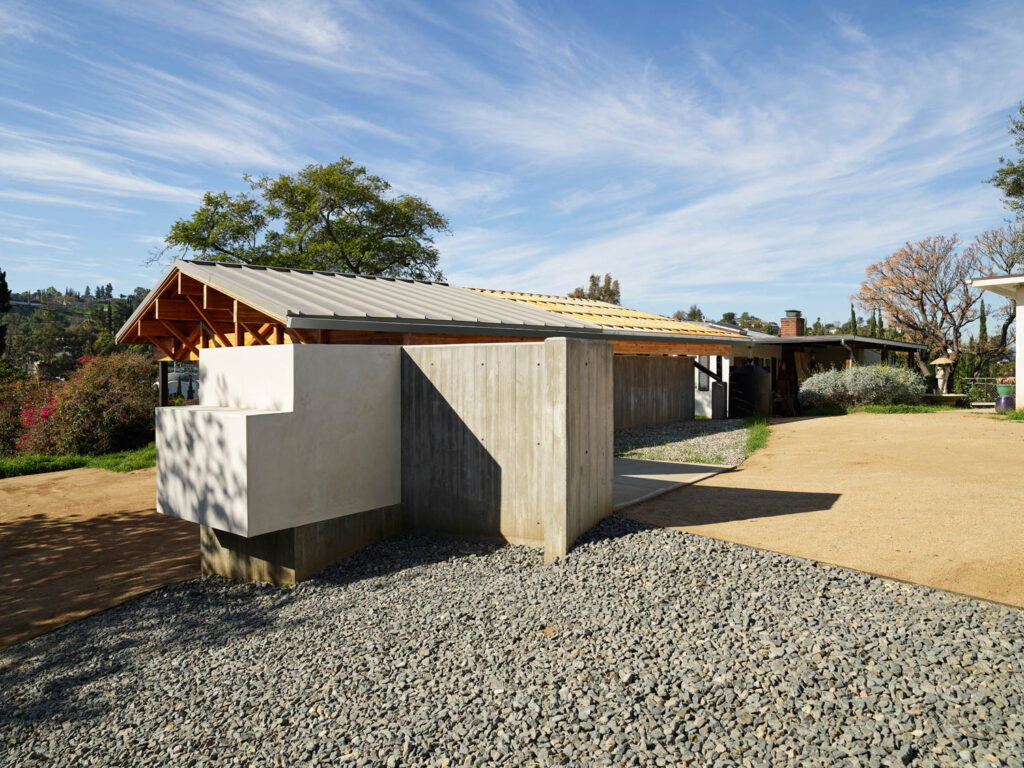 A modern building with a sloped metal roof, exposed wooden beams, and concrete walls, surrounded by gravel, dirt paths, and trees under a blue sky.