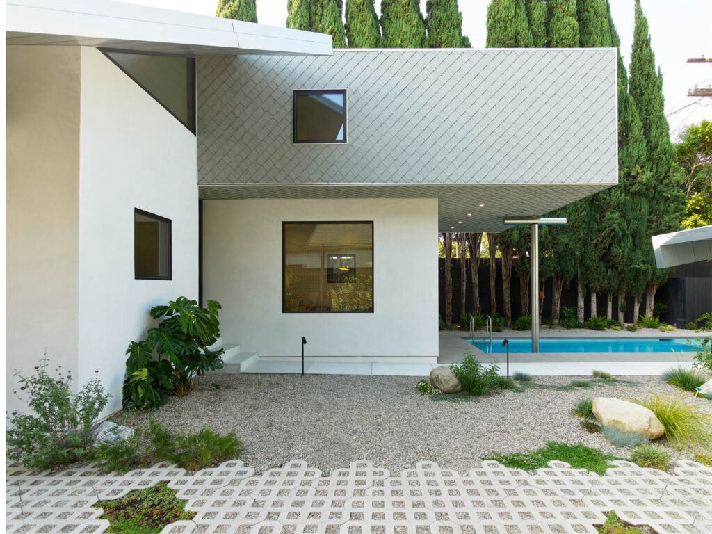 Modern house exterior with white walls, geometric windows, overhanging second floor, gravel yard, plants, and a swimming pool beside tall trees in the background.