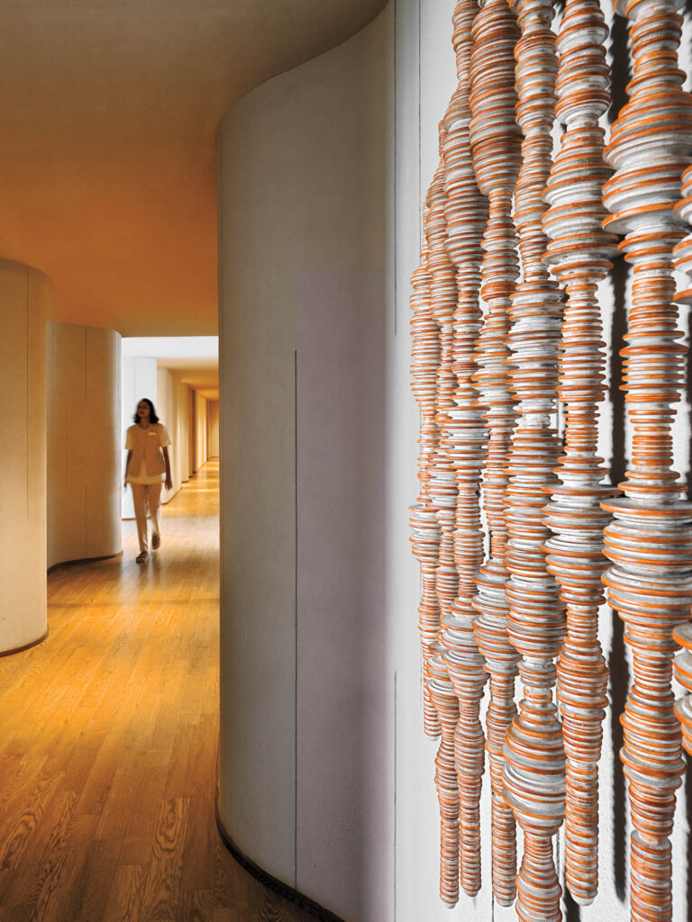 A person walks down a curved hallway with wooden floors; ceramic discs are artistically stacked and strung vertically along the wall in the foreground.