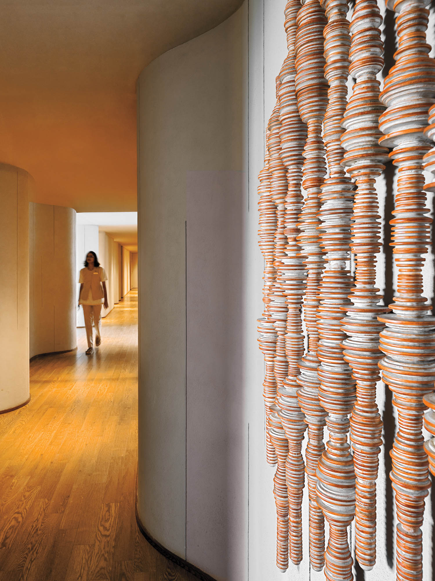 A person walks down a curved hallway with wooden floors; ceramic discs are artistically stacked and strung vertically along the wall in the foreground.