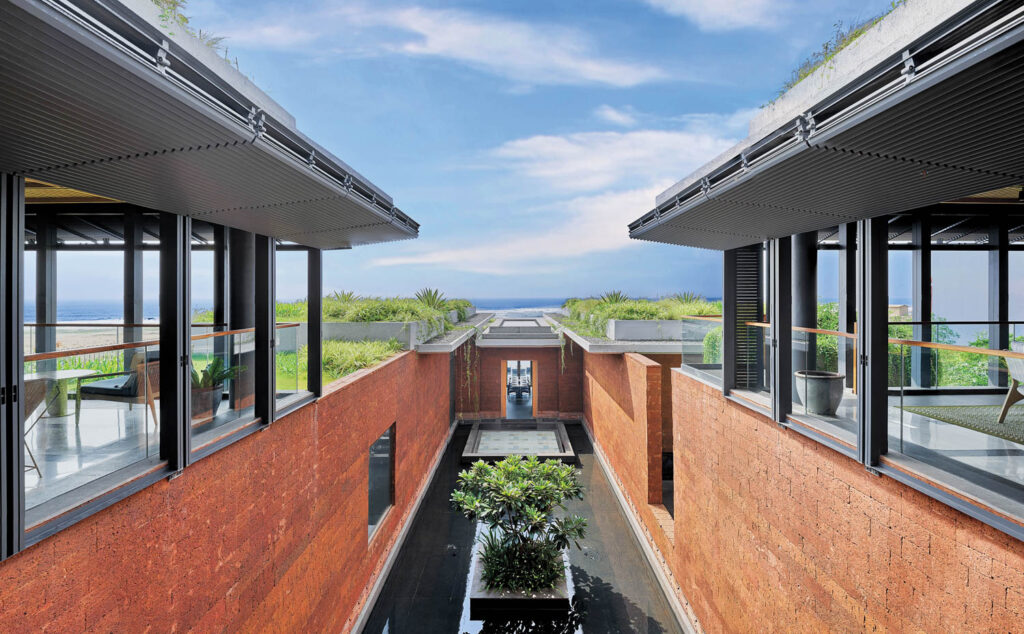 Modern building with red brick walls, large windows, and a central water feature with a tree, overlooking a garden and the ocean under a blue sky.