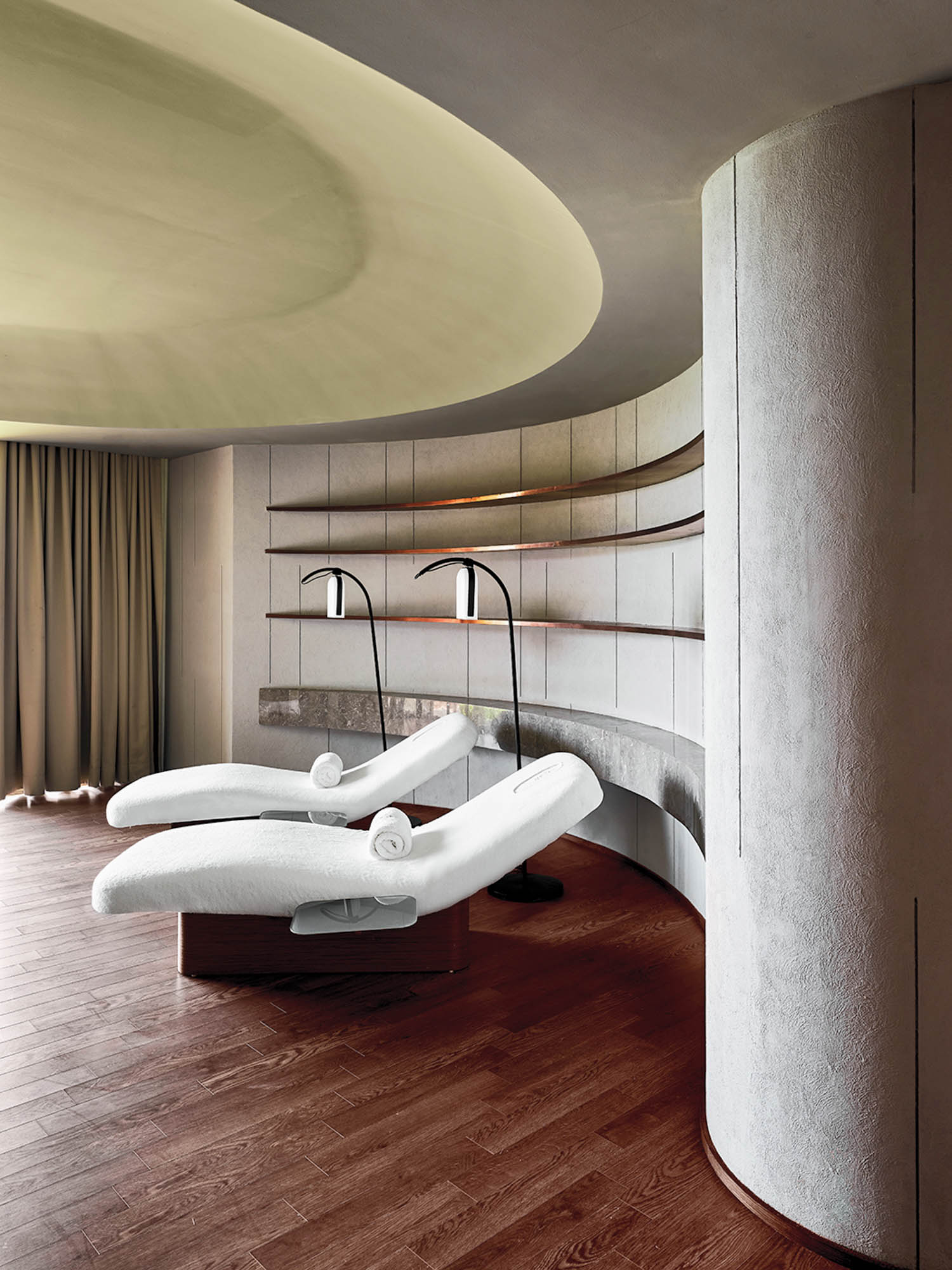 Two white massage beds with rolled towels are positioned in a modern, softly-lit spa room with curved walls, wooden floors, and tall arc floor lamps.