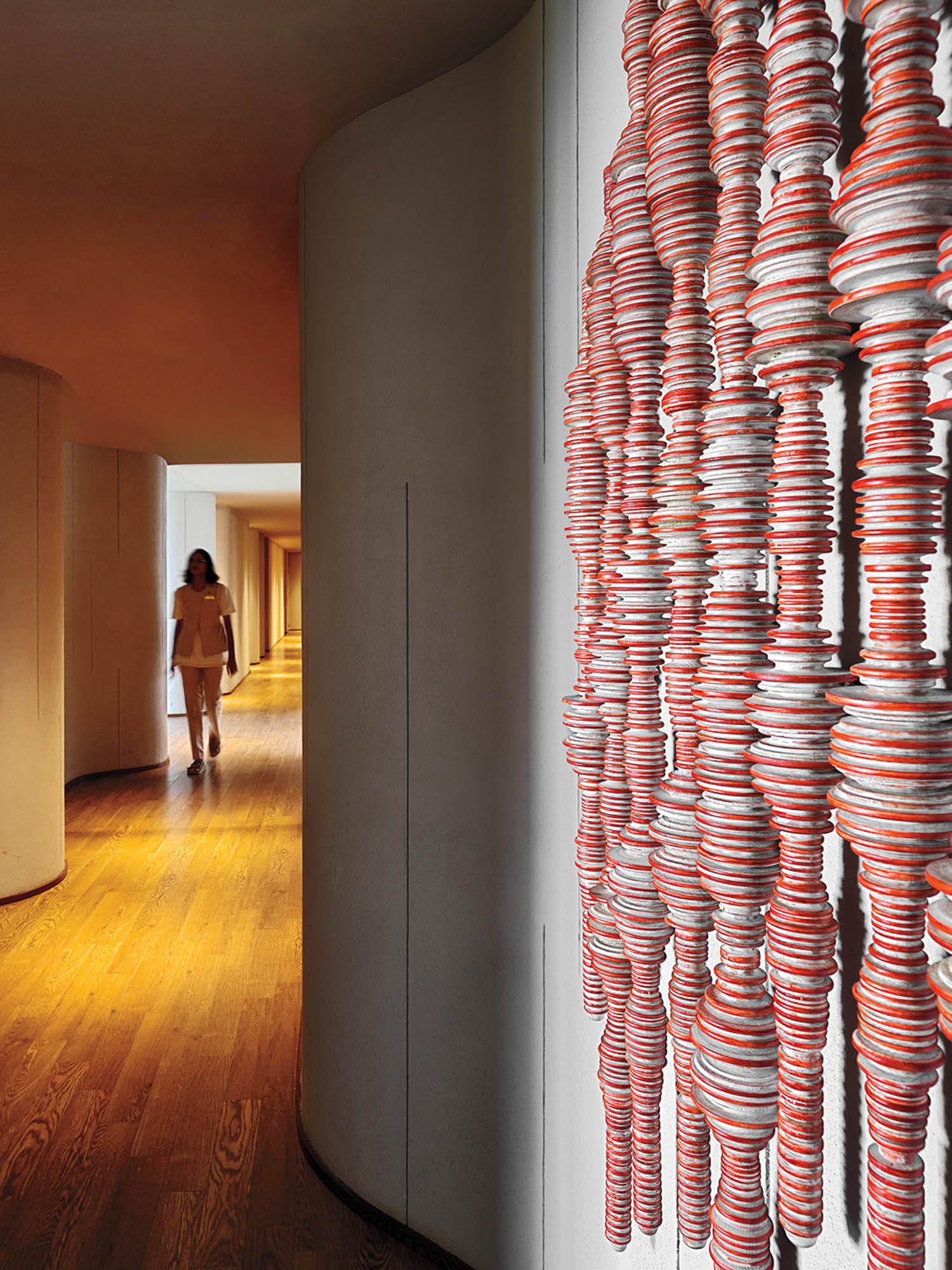 A person walks down a curved hallway with wooden floors; the foreground shows a vertical art installation made of stacked red and white discs.