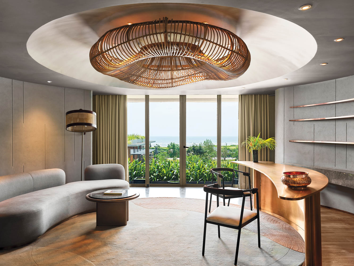 Modern living room with a curved sofa, round coffee table, black chair, built-in shelves, large windows with a garden and ocean view, and a woven ceiling light fixture.