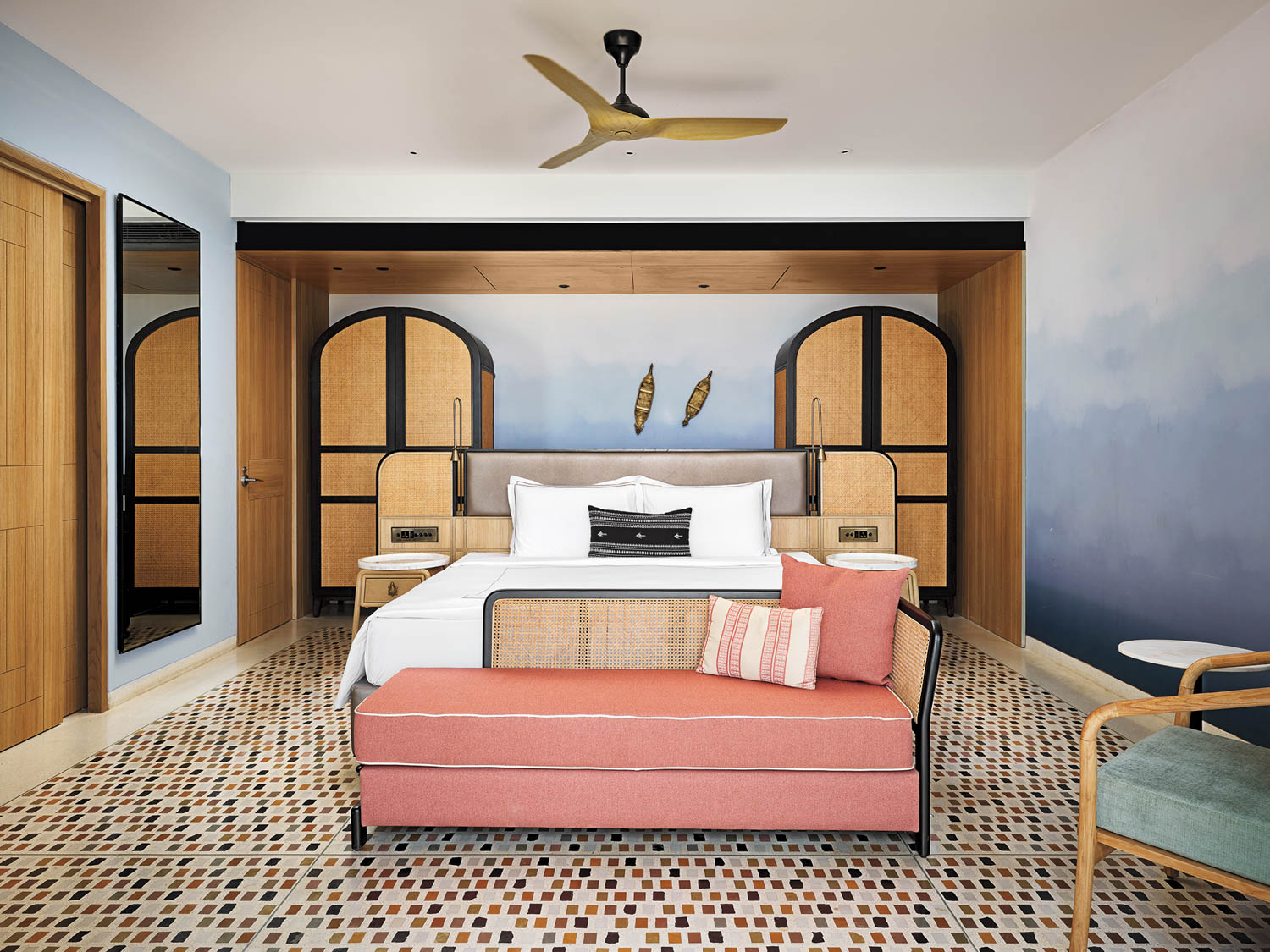 Modern hotel room with a double bed, wooden accents, mosaic tile floor, coral bench, two nightstands, wall art, and a ceiling fan.