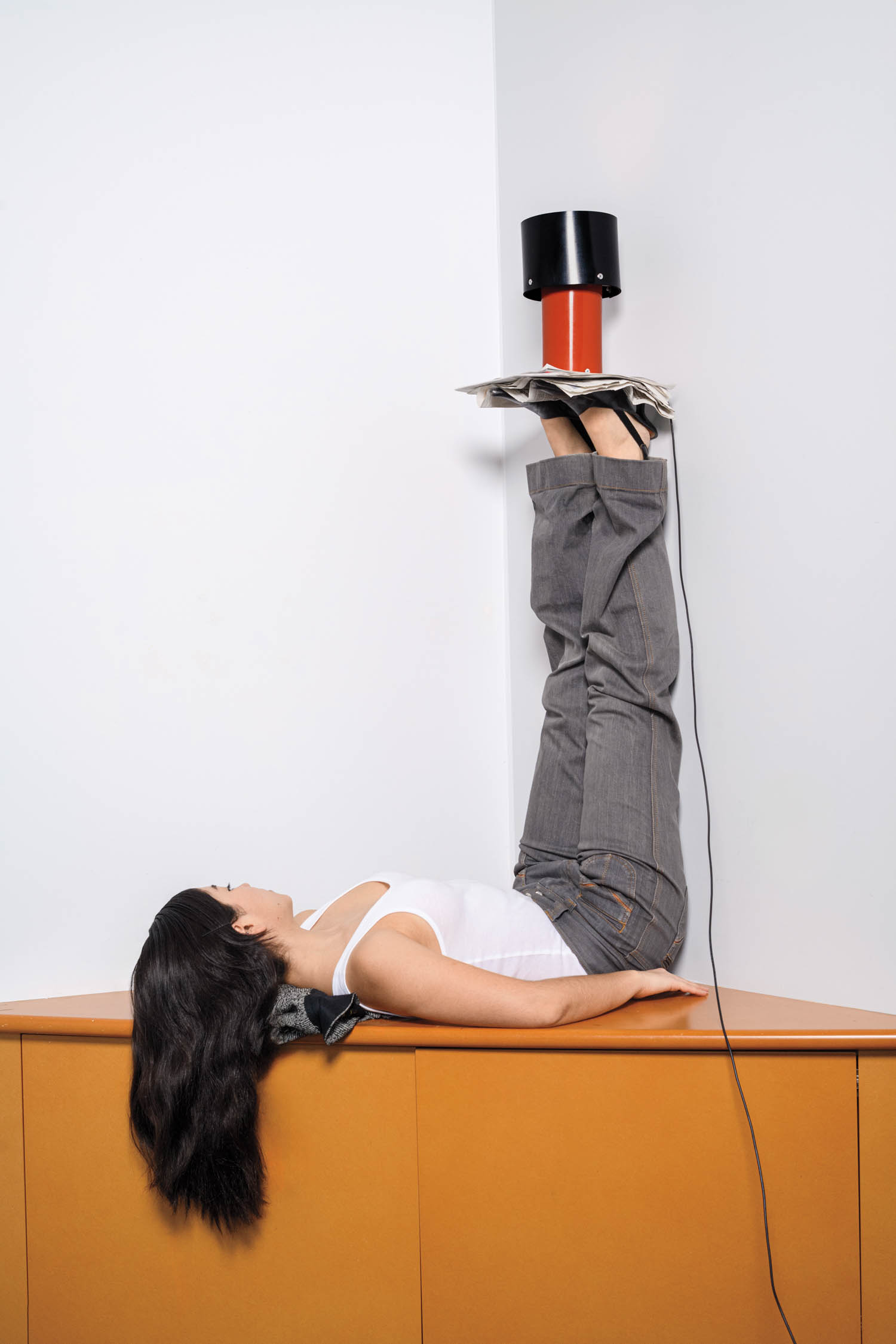 A person lies on a cabinet with legs raised vertically against a wall, feet under a black and red object, and head resting on a folded jacket.