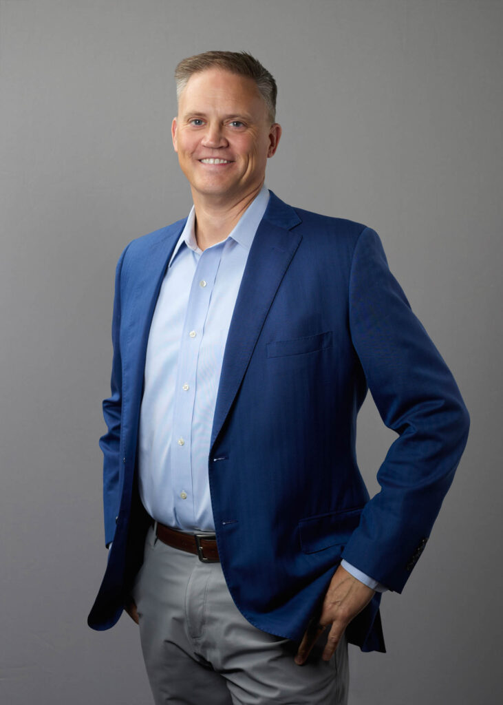 Man in a blue suit jacket and light blue shirt stands against a plain gray background, smiling with one hand in his pocket.