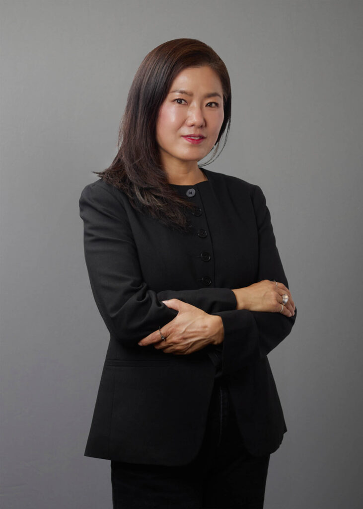 A woman with long dark hair in a black blazer stands with arms crossed against a plain gray background, looking at the camera.