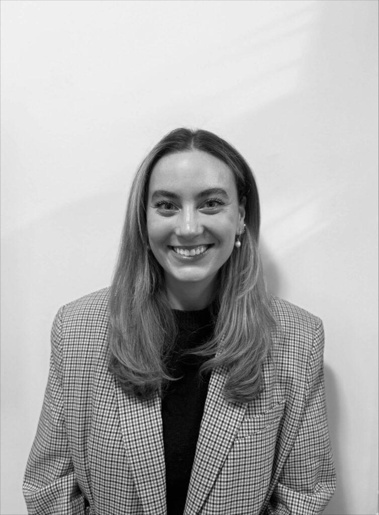 A woman with long hair and a checked blazer smiles at the camera, standing against a plain white background.