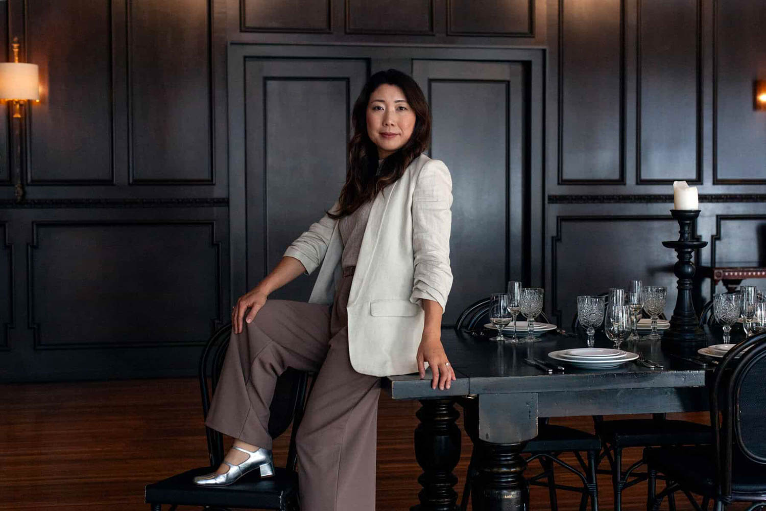 Woman in a light blazer and brown pants stands by a dining table set with glassware and plates in a dark-paneled room.