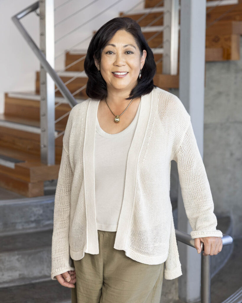 A woman with shoulder-length dark hair wearing a beige cardigan and light top stands indoors by a staircase, smiling at the camera.