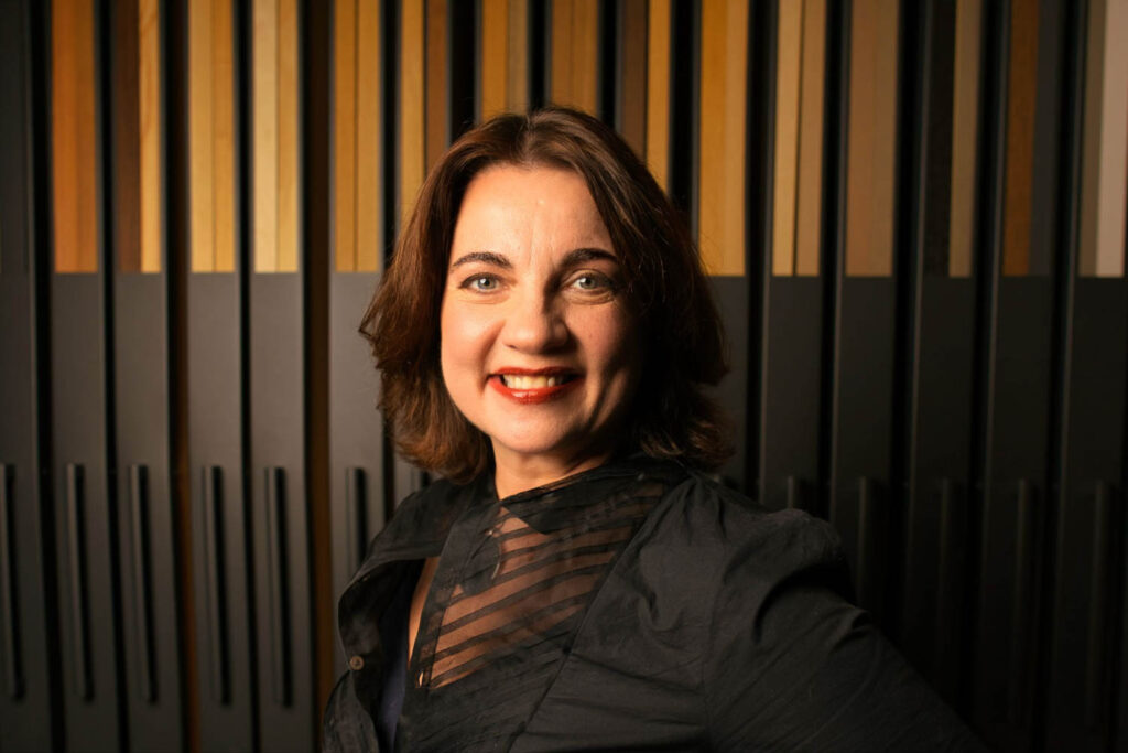 Woman with shoulder-length brown hair and a black blouse smiles at the camera, standing in front of a background with vertical black and wood-colored panels.