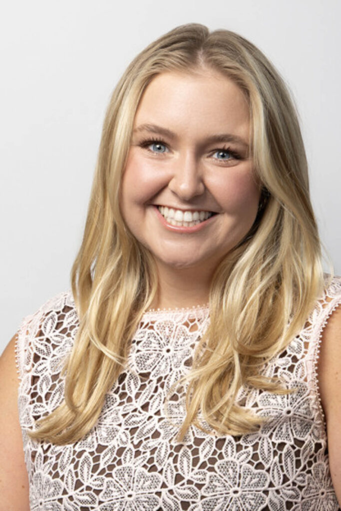 Woman with long blonde hair smiling at the camera, wearing a white lace top, in front of a plain light background.