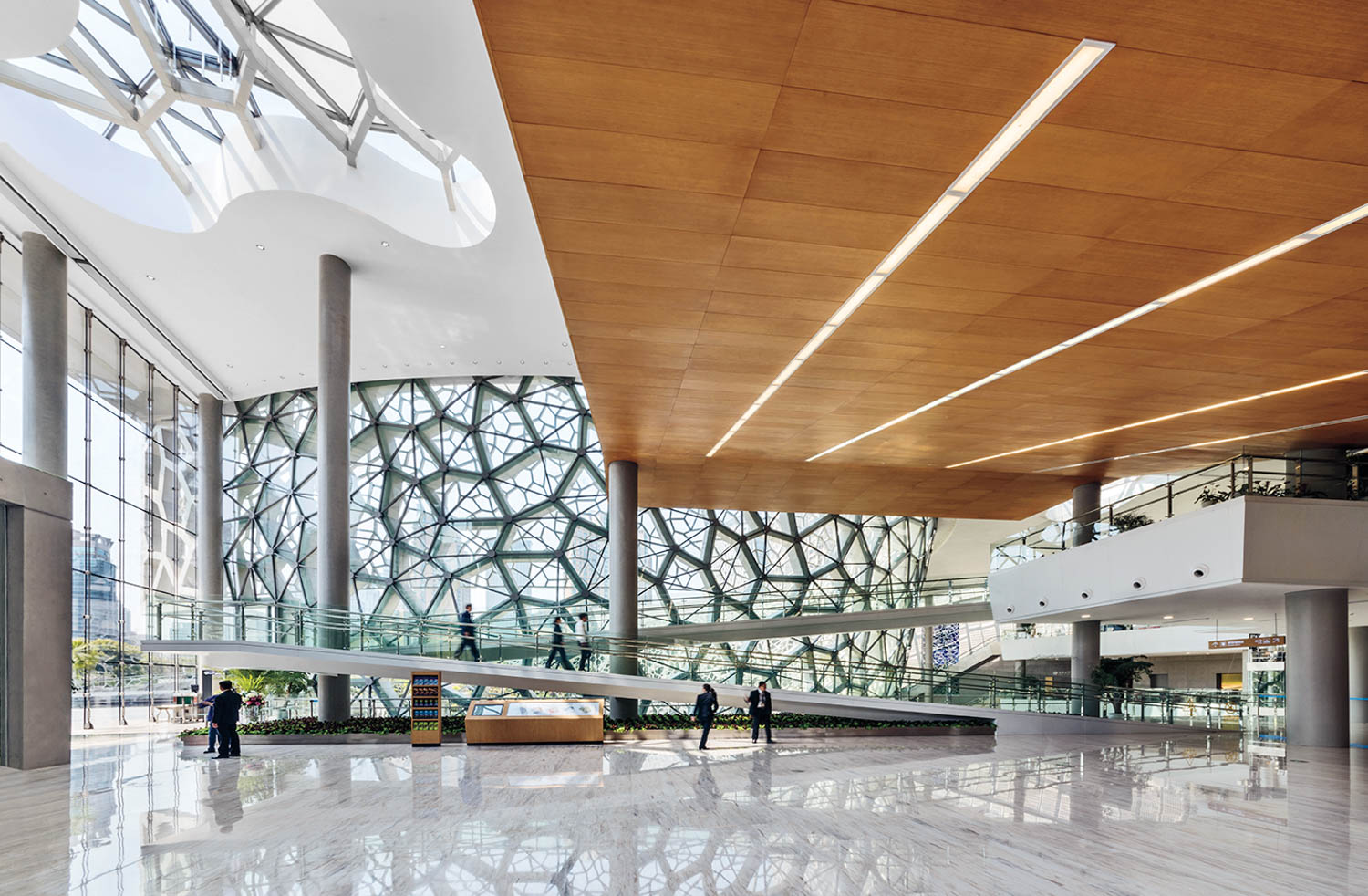 Spacious modern atrium with large glass walls, wooden ceiling panels, marble floors, and a gently sloping ramp with people walking.