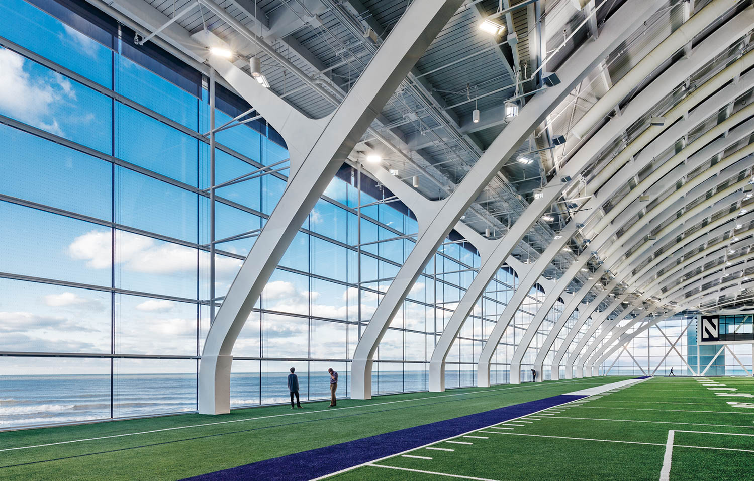 Two people stand on an indoor football field with large windows offering a view of the ocean and sky.