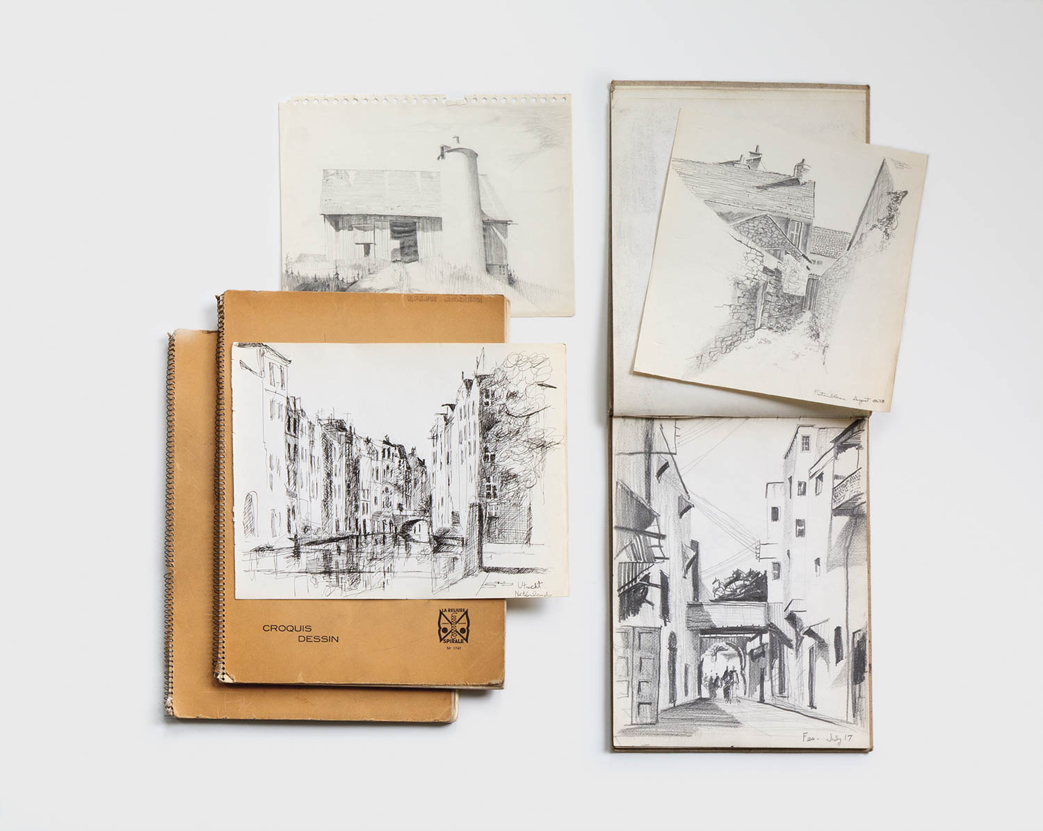 Five black-and-white pencil sketches of buildings and streets are arranged on a white surface, with two tan sketchbooks partially visible beneath the artwork.