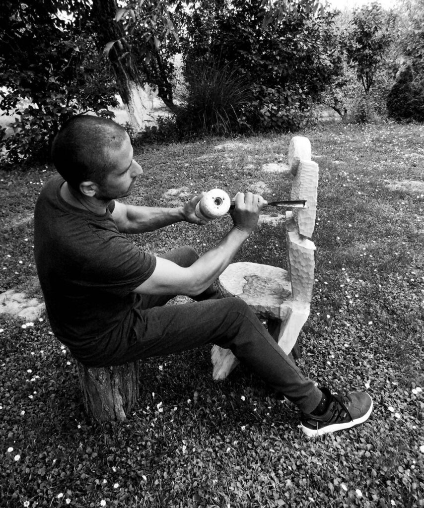 A man sits on a tree stump outdoors, sculpting a wooden chair with a mallet and chisel.