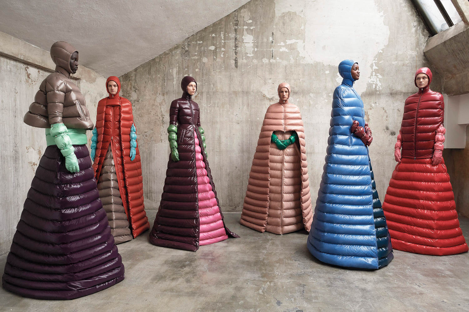 Six people stand in a concrete room wearing long, puffy, color-blocked coats with hoods and matching gloves, arranged in a semi-circle.