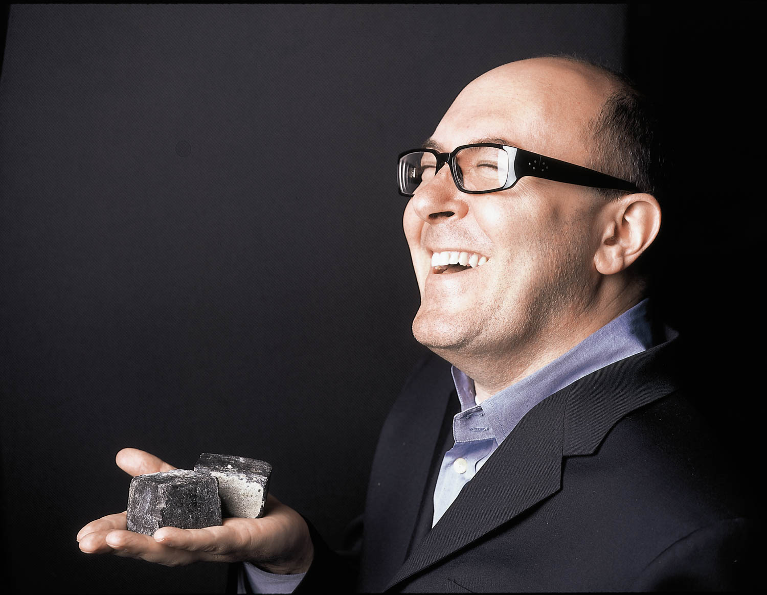 A smiling man in glasses and a suit holds several dark, square-shaped objects in his open palm against a black background.