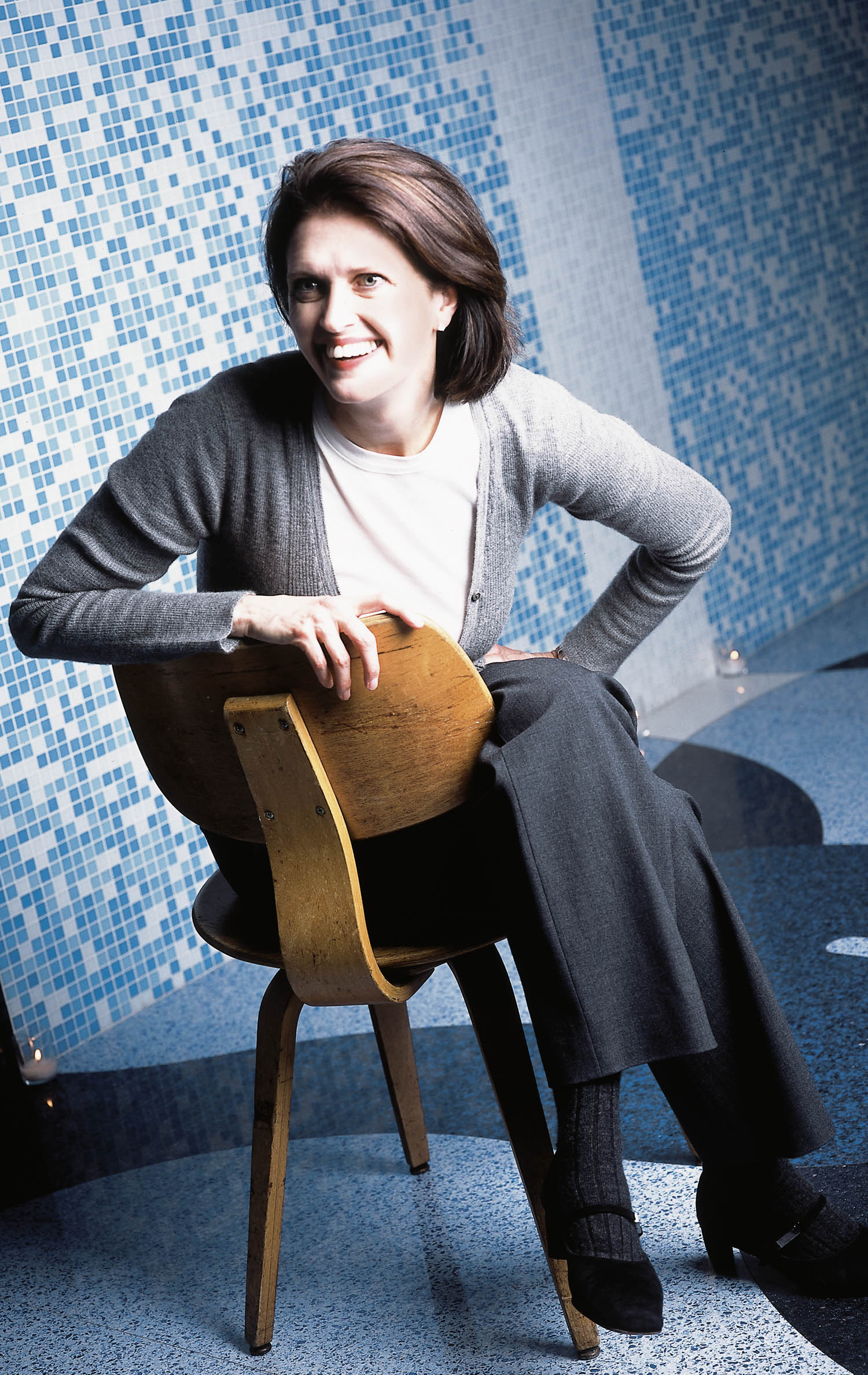Person with short brown hair sits sideways on a wooden chair, smiling, wearing a gray cardigan and dark pants. Blue and white mosaic tile wall in the background.