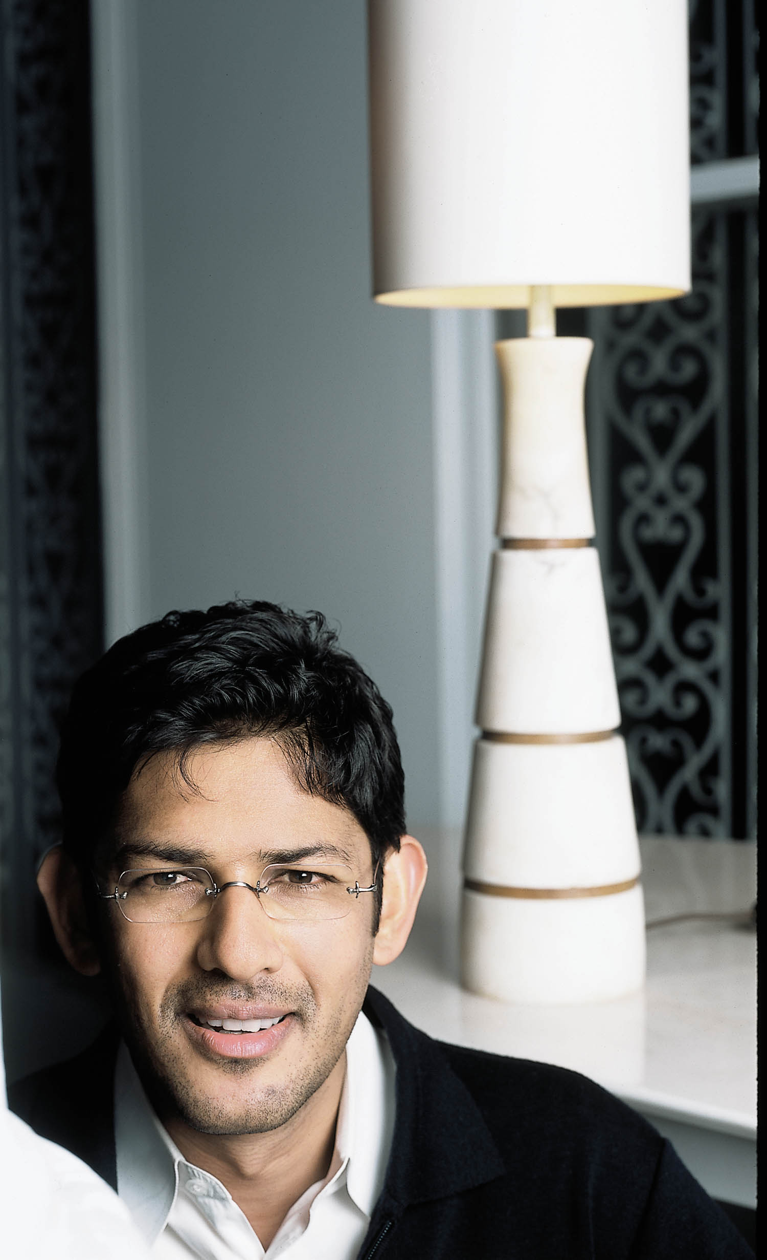 A man wearing glasses and a dark jacket sits indoors in front of a marble lamp with a white shade.