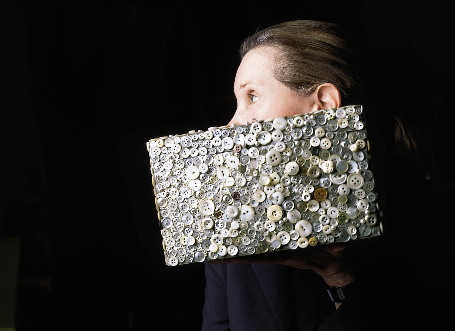 A person holds a rectangular object covered in assorted white buttons, partially obscuring their face, against a black background.
