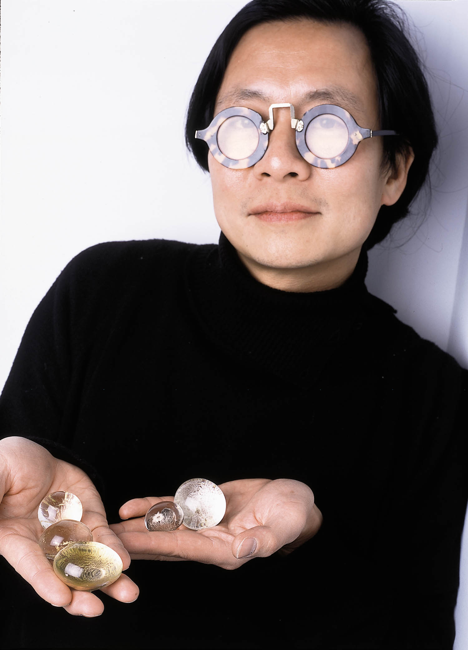 Person with round, reflective glasses and black turtleneck holds several round, translucent objects in open hands against a plain background.