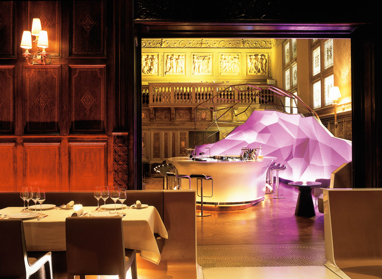 Elegant restaurant with set tables in the foreground; a modern, purple-lit bar with geometric design stands against ornate, classical walls and balcony in the background.