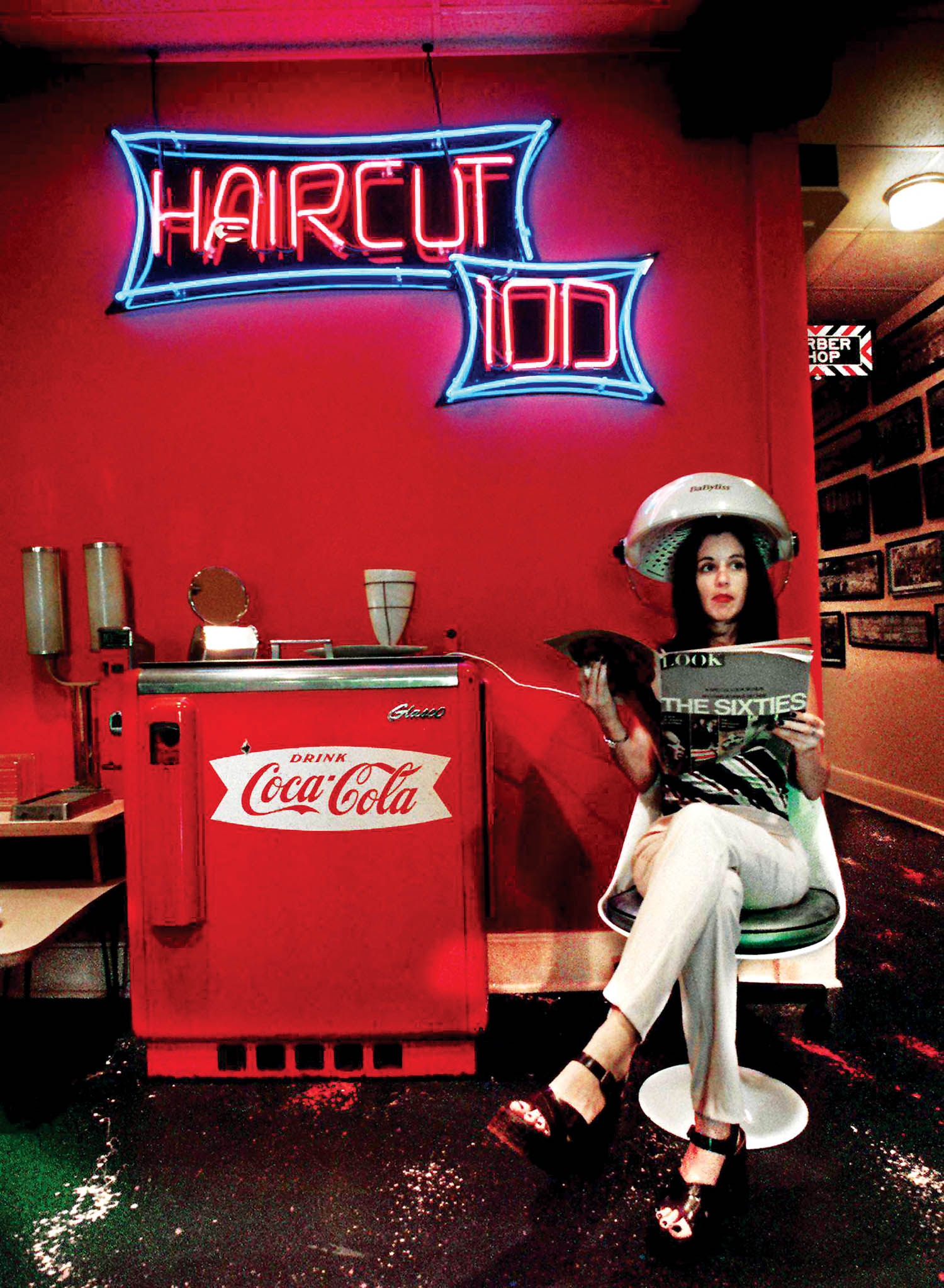 Woman sitting under a vintage hair dryer, reading a magazine in a retro salon with red walls, neon “HAIRCUT 100” sign, and a Coca-Cola machine.