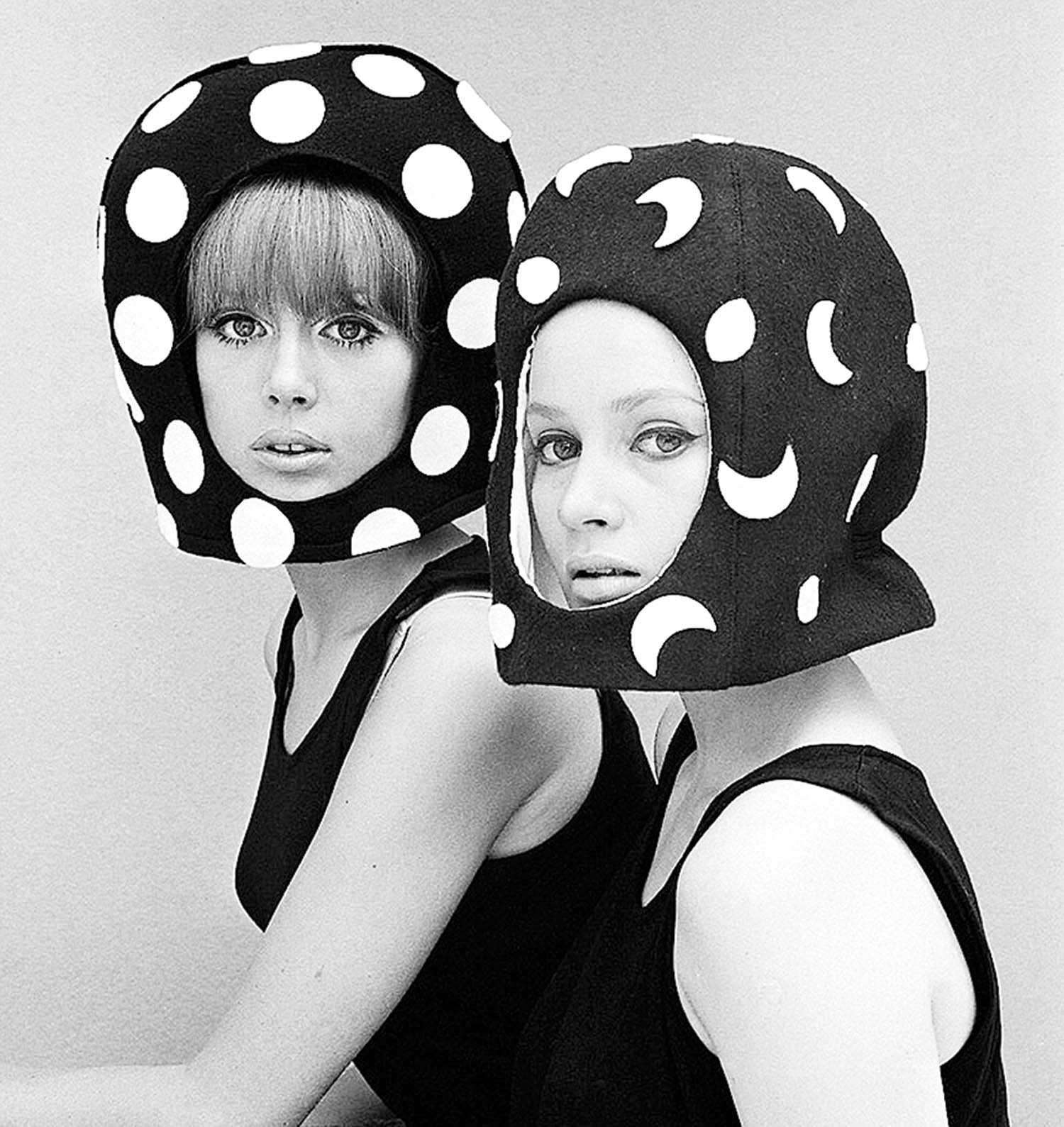 Two women in sleeveless black tops wear bold helmets, one with white polka dots and the other with white crescent shapes, posing against a plain background.