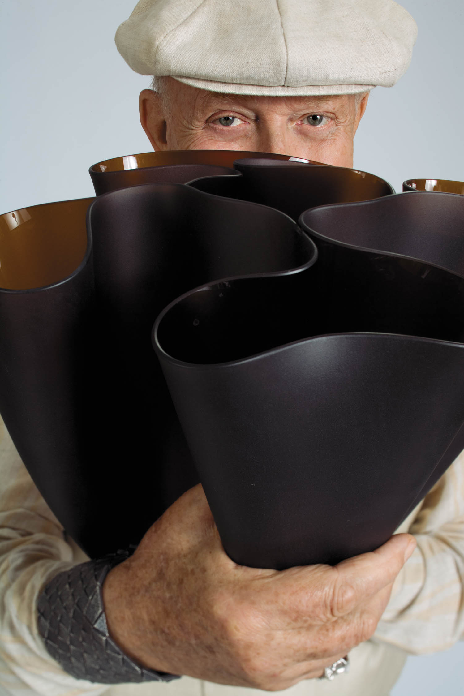 An older person in a light shirt and hat holds a cluster of large, dark, wavy-edged vases, partially obscuring their face.