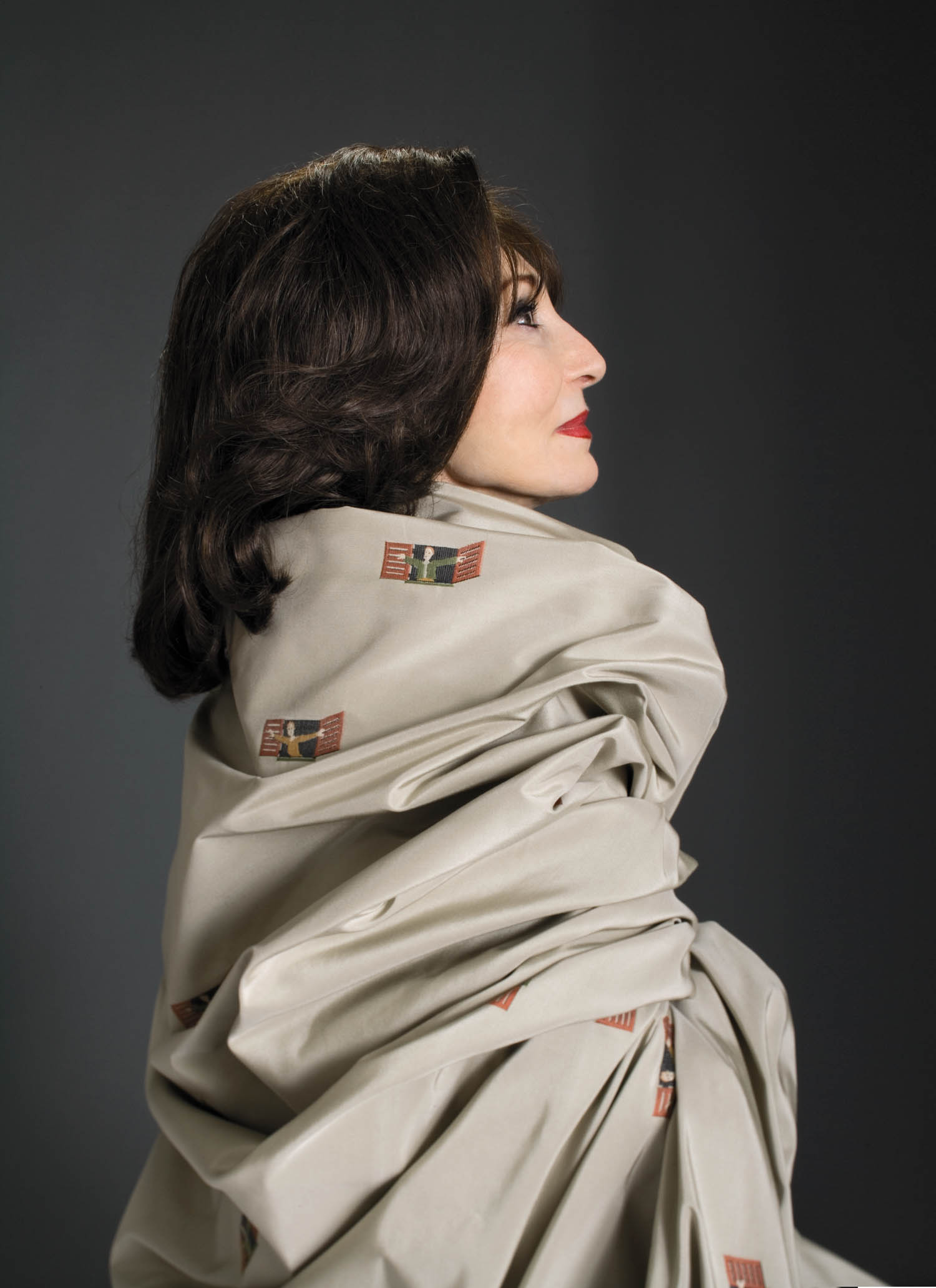 Woman with dark hair and red lipstick in profile, wrapped in a light-colored fabric decorated with small rectangular patterns, set against a plain dark background.