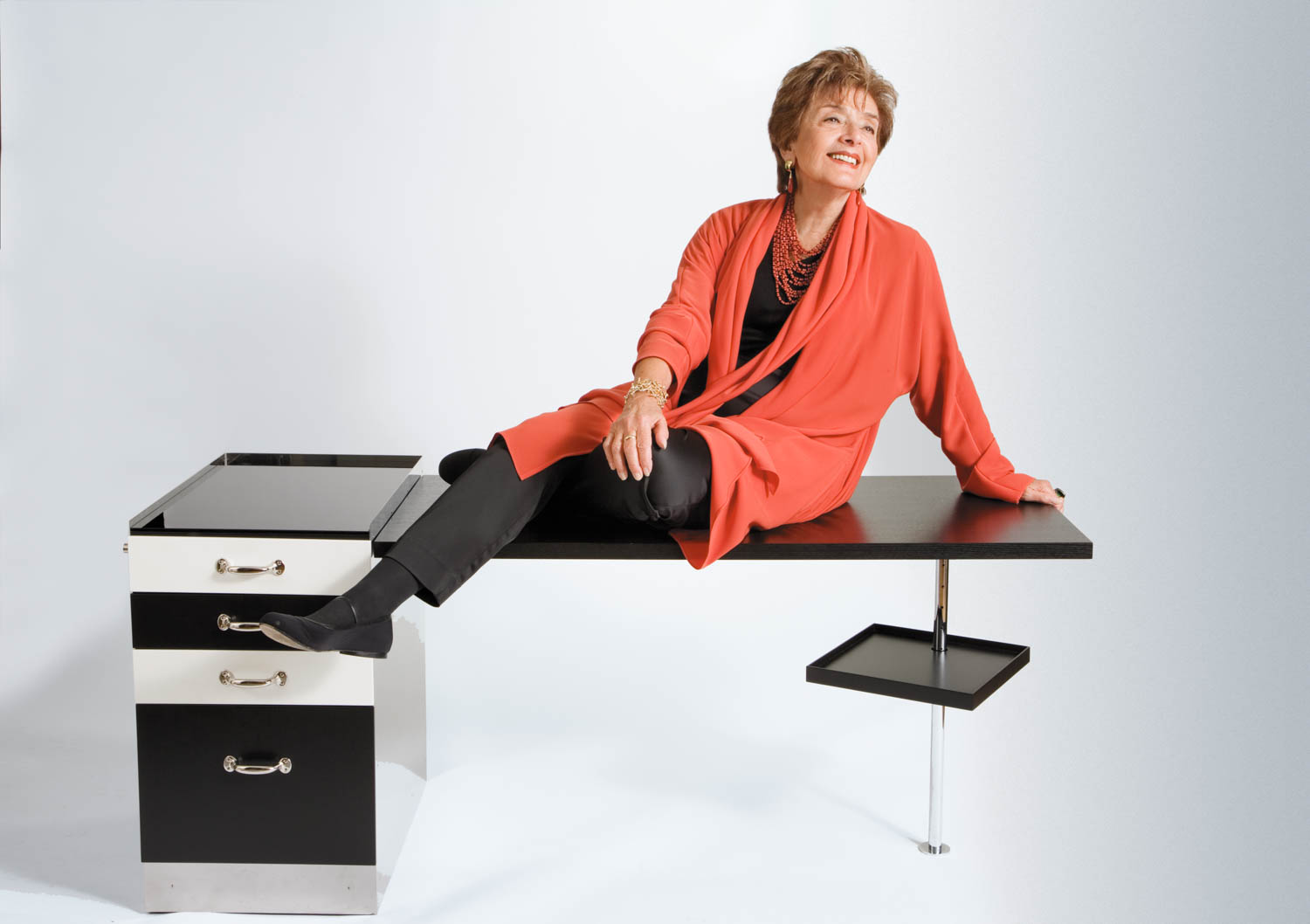 Woman in a red cardigan and black pants sits smiling on a modern black and white desk, with one leg crossed and her hand resting on the desk.