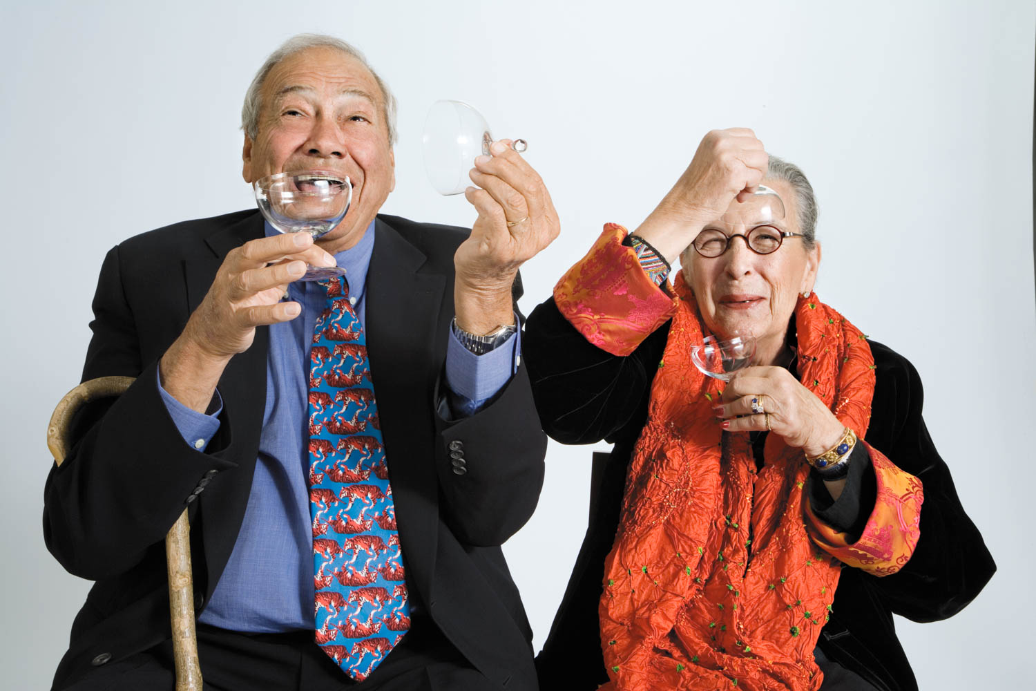 Two older adults sit side by side, smiling and playfully holding eyeglasses to their faces. One holds a cane, and both wear colorful clothing against a plain background.