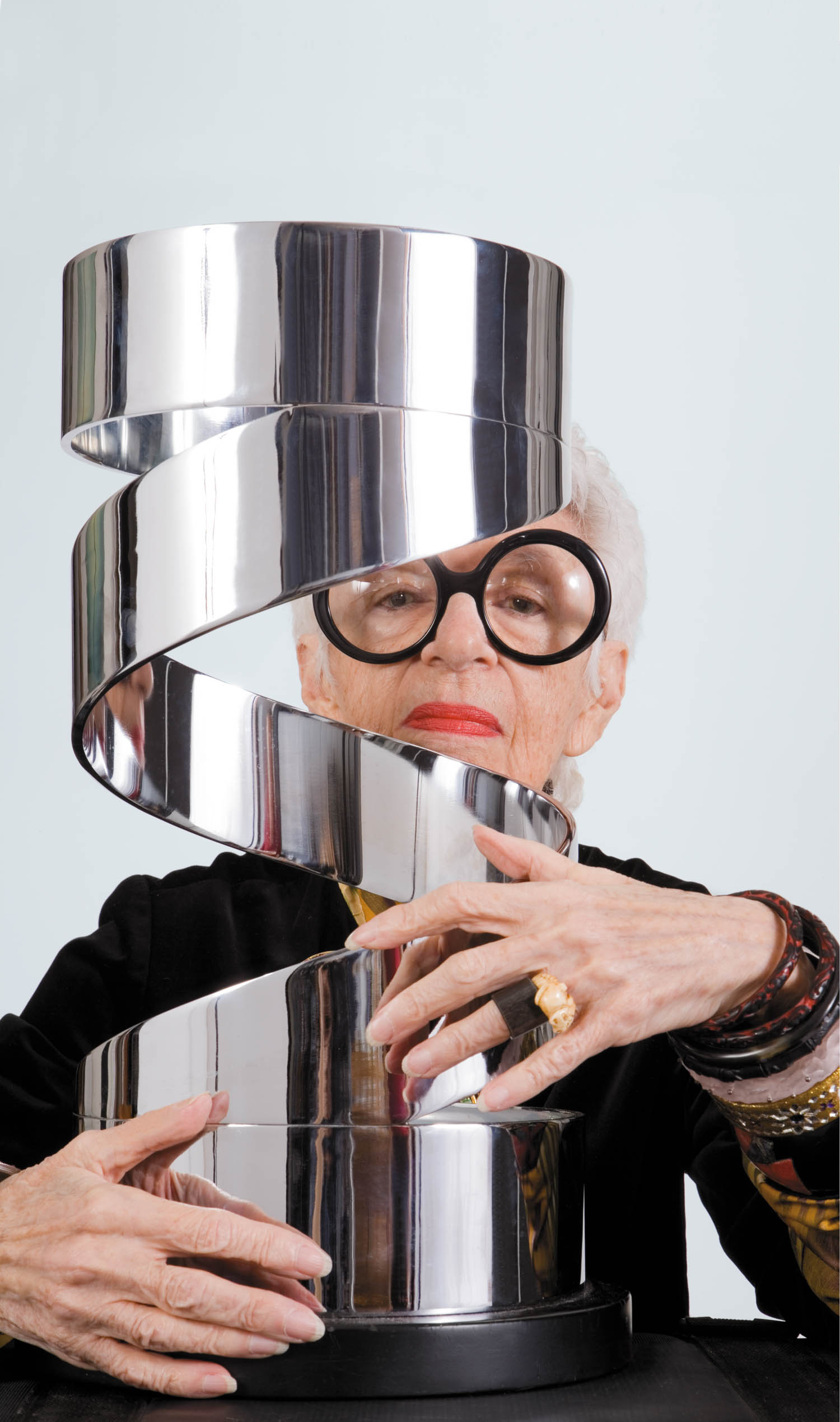 An older person with white hair and large round glasses holds a shiny, spiral-shaped metal sculpture against a plain light background.