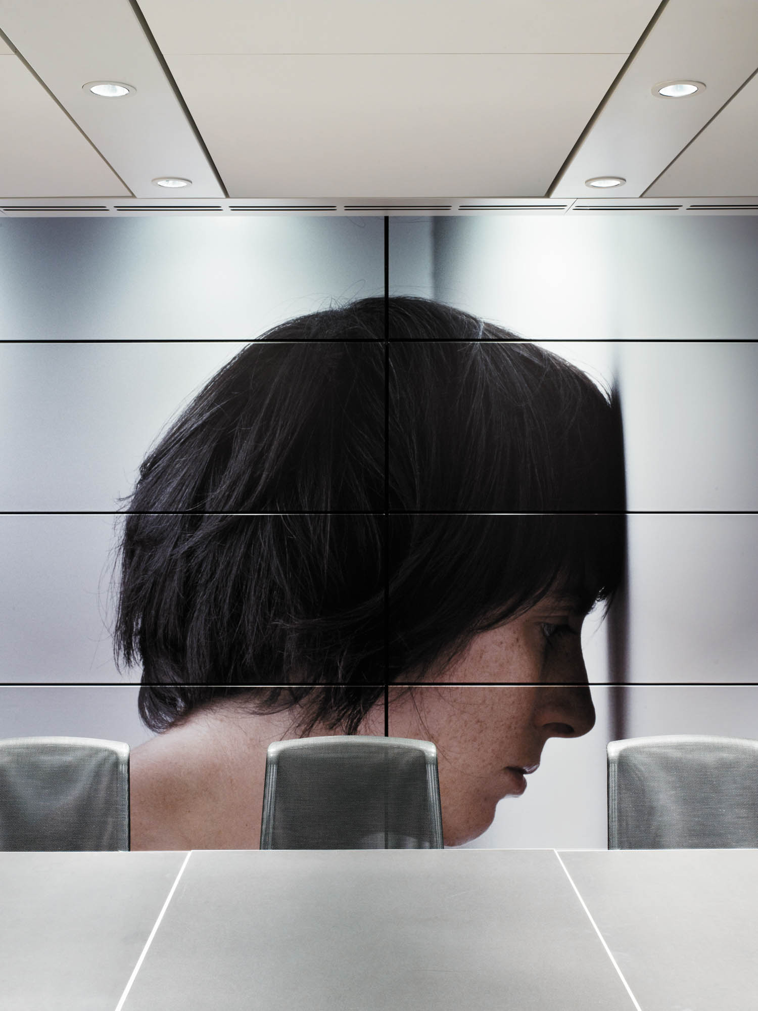 A large wall mural shows a close-up side profile of a person with short dark hair, their forehead touching the wall, behind a row of empty conference chairs.