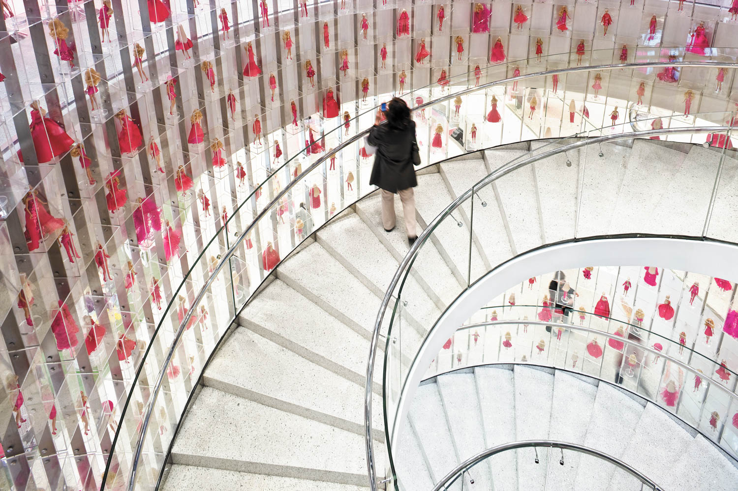 Person walking down a curved staircase surrounded by transparent walls with small red dress ornaments hanging inside.