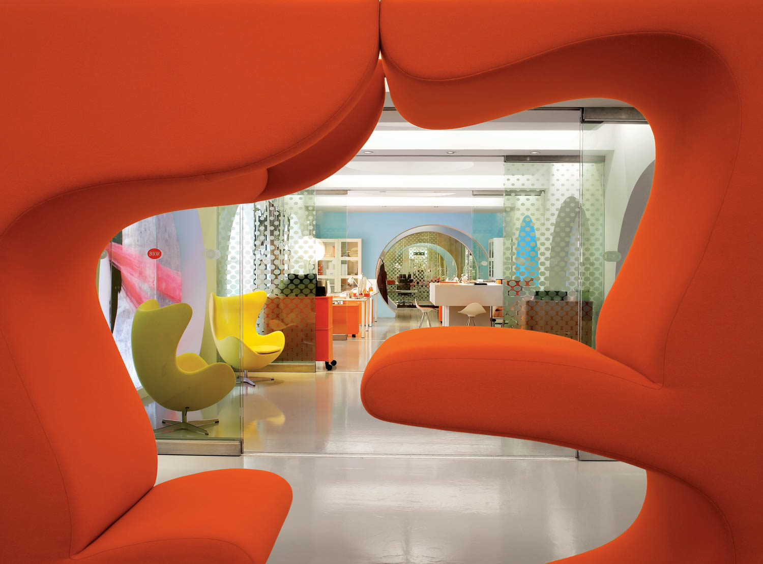View through two large orange sculptural chairs to a modern office space with yellow chairs, glass walls, and eclectic decor.