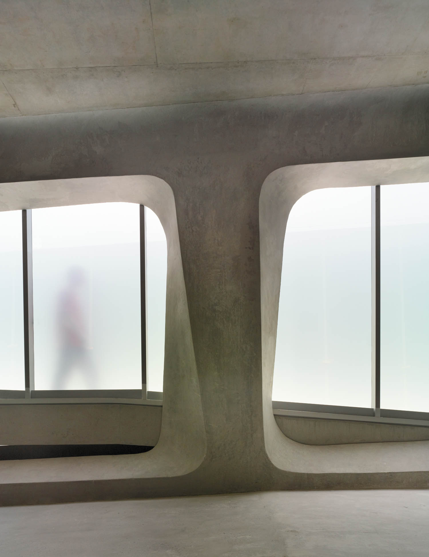 A blurred person walks behind frosted glass windows in a modern, minimalist concrete interior with geometric openings.